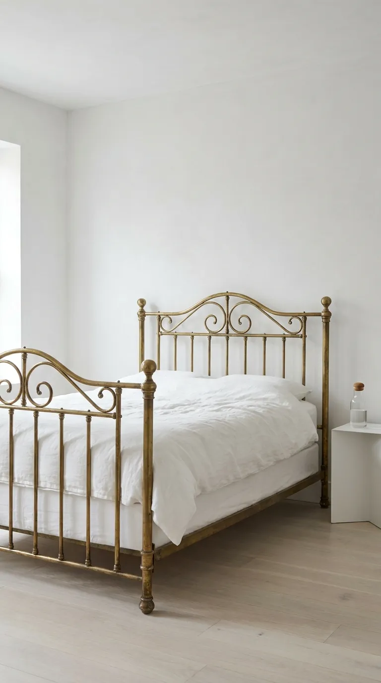 Editorial photograph of an ornate antique brass bed frame with delicate scrollwork and softly tarnished patina, set within an entirely white modern bedroom featuring smooth plaster walls, white oak flooring, and simple white linen bedding with no additional color accents. A single small glass carafe of water sits on a minimalist white nightstand beside the bed. Soft even daylight fills the room from a large unseen window, allowing the brass frame's warm tone and texture to stand out as the sole focal point. Clean, gallery-like composition, no people, no text overlays.